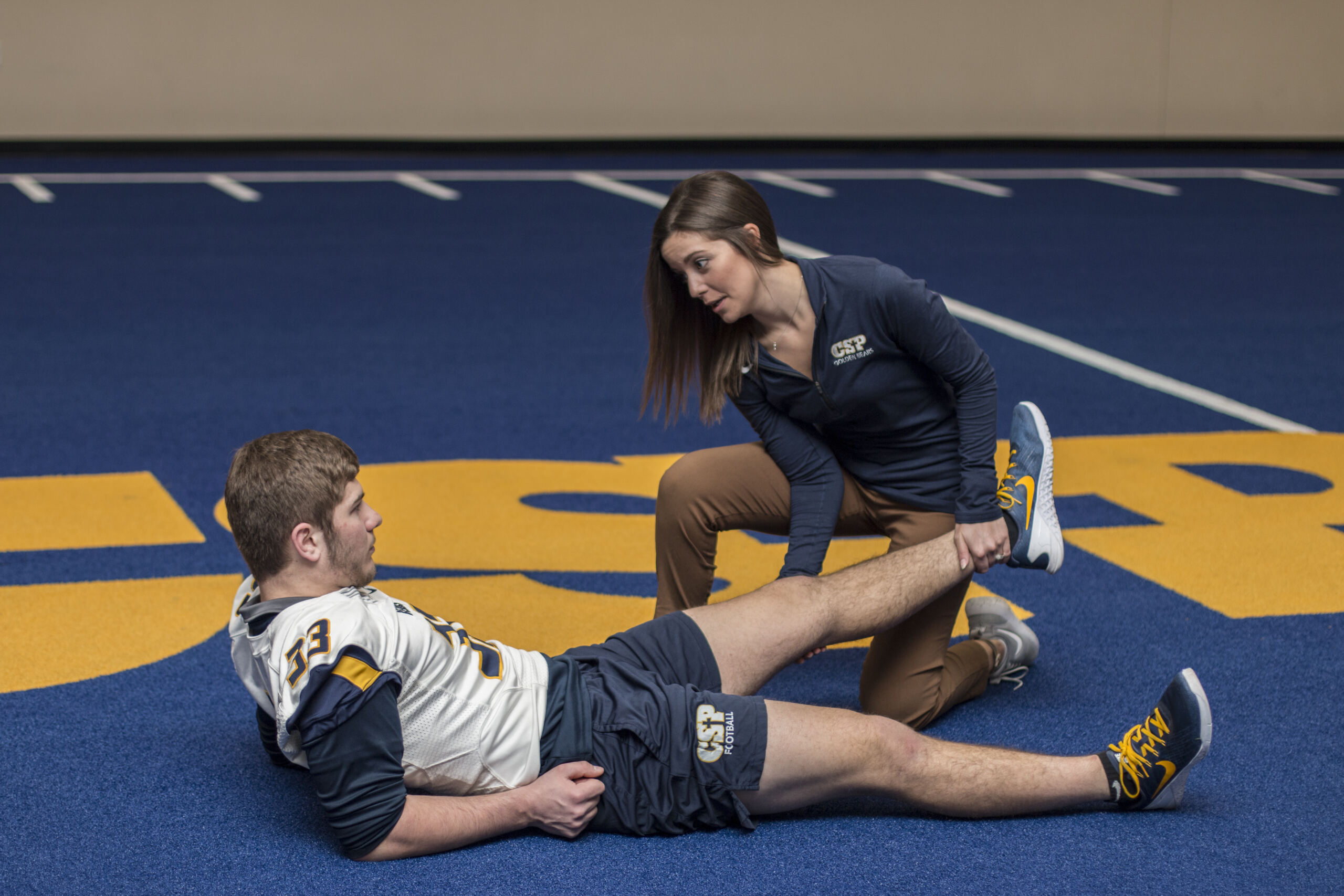 A female exercise physiologist assisting a young athlete with a leg stretch during rehabilitation on an indoor sports field, illustrating clinical and athletic care in exercise physiology
