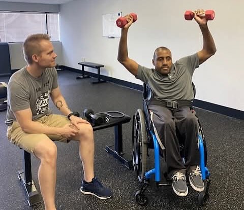 Dr. Mark Fleming watches on at eye level as a client with spina bifida and scoliosis does dumbbell shoulder presses to ensure the lift is being done properly and safely.